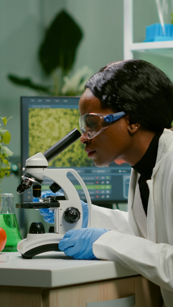 a woman wearing goggles and white coat looking through a microscope